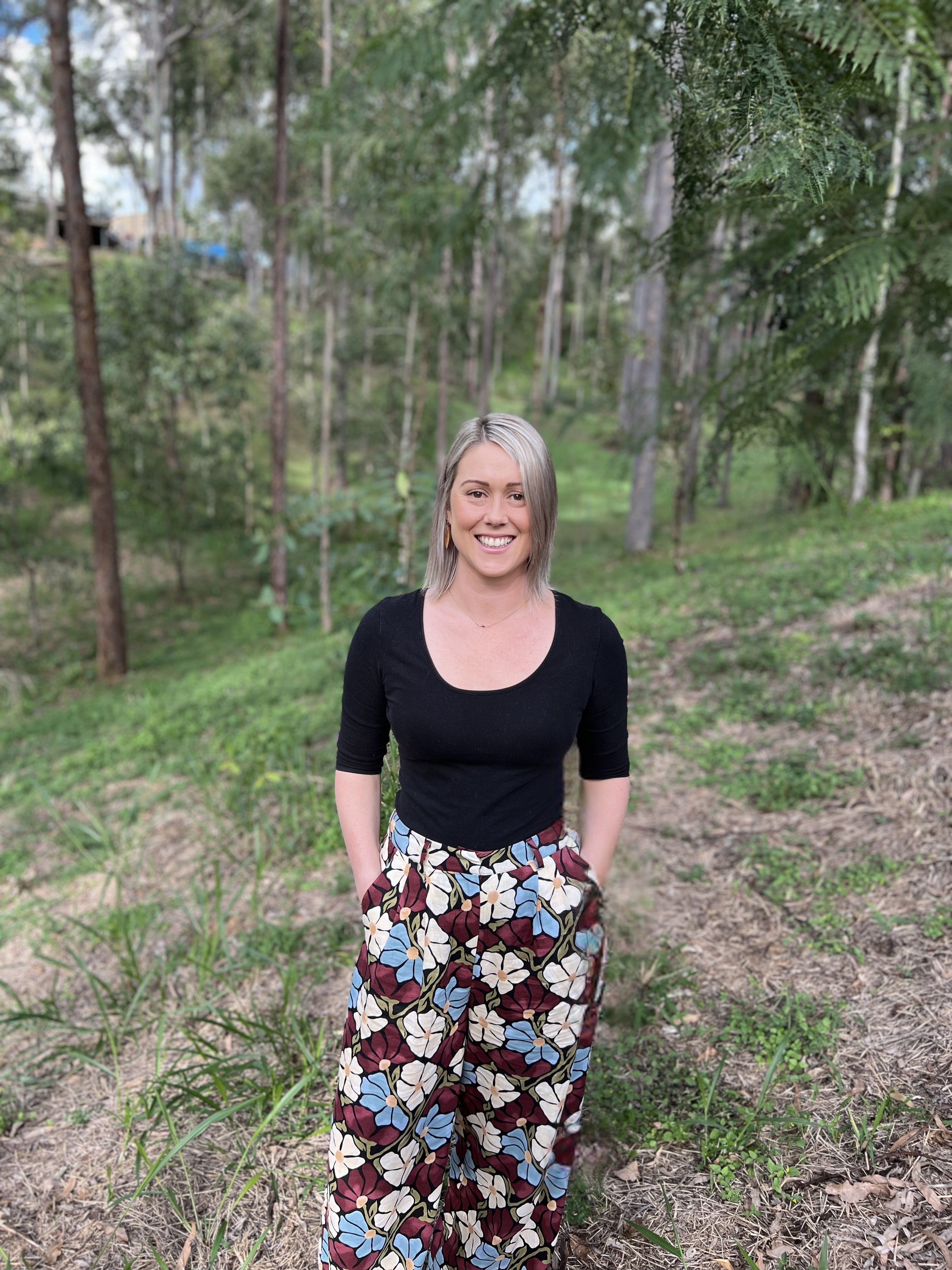 Woman standing in a forest wearing a black top and patterned pants.