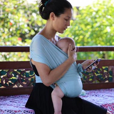 Woman wearing a baby in a sling and using a phone on a balcony with greenery in the background on the sunshine coast